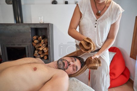Téléchargez les photos : Crop femme thérapeute en lin blanc t-shirt pétrissant le cou du client masculin barbu avec les yeux fermés lors de la séance de massage dans la chambre avec cheminée - en image libre de droit