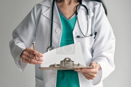 Téléchargez les photos : Cultiver médecin femme méconnaissable en robe médicale avec crayon et papier sur presse-papiers sur fond clair - en image libre de droit