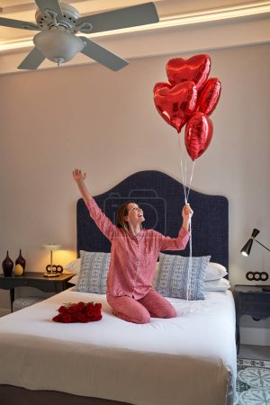 Foto de Feliz joven hembra en pijama sentada en la cama con ramo de rosas y levantando las manos con manojo de globos de papel de aluminio rojo mientras regocija regalo en la habitación de hotel - Imagen libre de derechos