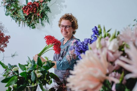 Foto de Florista femenina adulta positiva con pelo rizado rubio corto y gafas de pie con ramo de flores rojas rodeadas de varias plantas en la tienda de flores - Imagen libre de derechos
