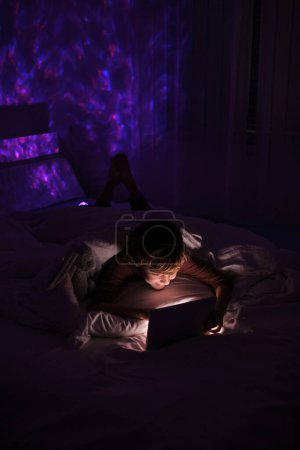 Téléchargez les photos : Comprimé de navigation garçon insomnie tout en étant couché sur le lit sous couverture dans la chambre à coucher avec une lumière tamisée la nuit à la maison - en image libre de droit