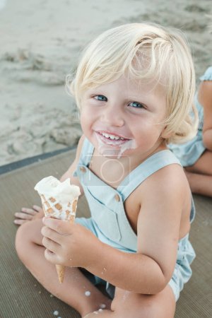 Foto de Alto ángulo de niño alegre con helado sonriendo y mirando a la cámara mientras descansa en la playa - Imagen libre de derechos
