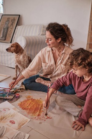 Photo for High angle of concentrated kid and mom painting with gouache while sitting on floor at home near Spaniel dog - Royalty Free Image