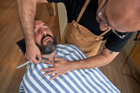 Foto de Alto ángulo de barba barbuda barba de corte macho del cliente con hoja recta mientras trabaja en la barbería - Imagen libre de derechos