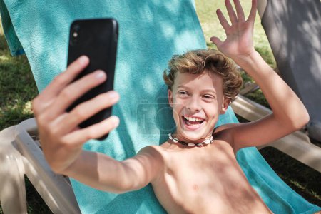 Photo for Happy boy with blond hair sitting on blue blanket and looking at screen of mobile phone while laughing with opened mouth and saying hello with raised hand fingers - Royalty Free Image