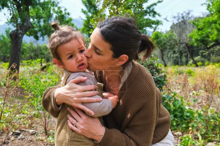 Téléchargez les photos : Positif jeune mère ethnique étreignant et embrassant la joue mignonne fille doucement dans le parc d'été verdoyant - en image libre de droit