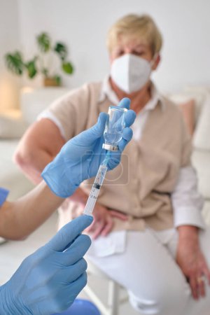 Photo for Crop anonymous physician in sterile gloves with injector and COVID 19 vaccine in bottle against senior female patient in house - Royalty Free Image