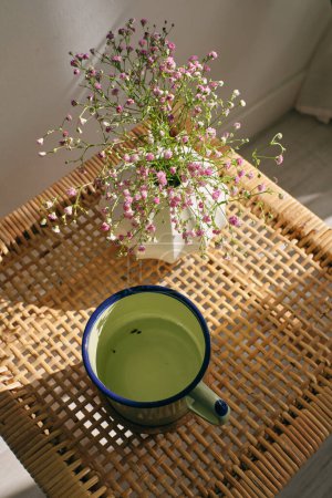Photo for From above of iron mug with aromatic green tea and blooming gypsophila flowers in vase placed on wicker table in cozy light room - Royalty Free Image