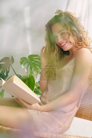 Téléchargez les photos : Portrait de jeune femme souriante en robe assise sur la chaise et le livre de lecture tout en se reposant dans une chambre confortable - en image libre de droit