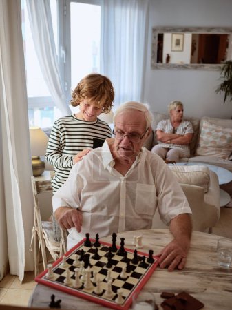 Foto de Niño positivo en ropa casual de pie cerca enfocado abuelo jugando ajedrez juego en la mesa en la sala de estar de luz en casa - Imagen libre de derechos