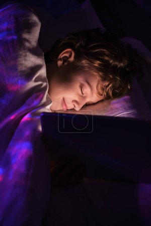 Foto de Lindo niño durmiendo cerca de la tableta mientras está acostado en la cama debajo de la manta en el dormitorio con iluminación tenue por la noche en el apartamento - Imagen libre de derechos