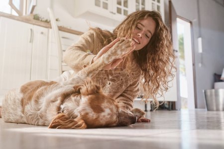 Téléchargez les photos : Niveau du sol de la jeune femme souriante avec de longs cheveux bouclés donnant haute cinq à chien mignon tout en jouant ensemble sur le sol de la cuisine - en image libre de droit