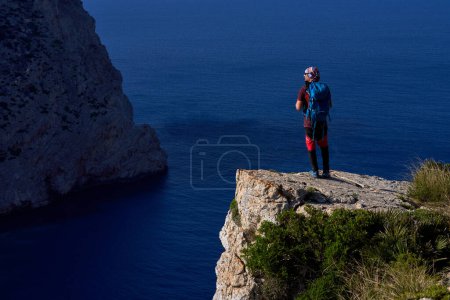 Téléchargez les photos : Vue arrière pleine longueur de l'homme avec sac à dos debout sur une falaise rocheuse et regardant loin contre l'océan par une journée ensoleillée - en image libre de droit