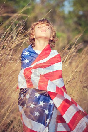 Téléchargez les photos : Rêveur préadolescent garçon roulé dans les étoiles nationales et rayures drapeau américain debout avec les yeux fermés et la tête pliée en arrière tout en passant du temps dans le champ - en image libre de droit