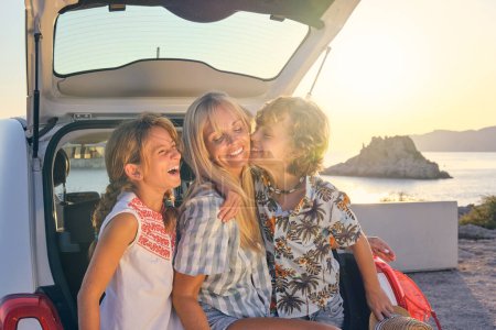 Téléchargez les photos : Rire frères et sœurs en vêtements d'été embrassant mère souriante tout en étant assis ensemble sur le coffre de la voiture sur le bord de la mer dans le dos éclairé - en image libre de droit