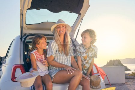 Foto de Madre con dos hijos en sombreros de paja y ropa de verano sentada en el maletero de un coche blanco durante el amanecer con el mar al fondo - Imagen libre de derechos
