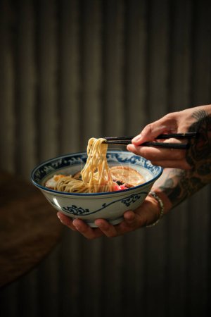 Foto de Cultivo persona anónima con palillos comiendo deliciosos fideos de sopa de ramen japonés de cuenco en habitación oscura en el restaurante - Imagen libre de derechos