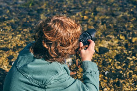 Téléchargez les photos : Vue de dessus latérale d'un garçon méconnaissable prenant une photo de l'eau transparente avec du caillou assis sur la côte dans une nature ensoleillée - en image libre de droit