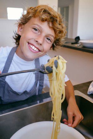 Photo for Content child looking at camera with cooked pasta on spatula above sink at home in daytime - Royalty Free Image