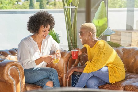 Foto de Mujeres multirraciales positivas con tazas de té caliente riendo mientras están sentadas en un sofá en una habitación soleada y conversando - Imagen libre de derechos