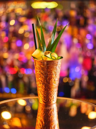 Téléchargez les photos : Tasse en cuivre avec cocktail de mule de Moscou alcoolisé décoré de concombre et de feuilles vertes servi dans un bar moderne sur fond flou - en image libre de droit