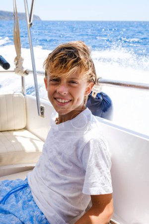 Téléchargez les photos : Joyeux garçon préadolescent aux cheveux blonds mouillés en t-shirt blanc souriant et regardant la caméra alors qu'il était assis dans un bateau moderne pendant la croisière en mer par une journée ensoleillée - en image libre de droit