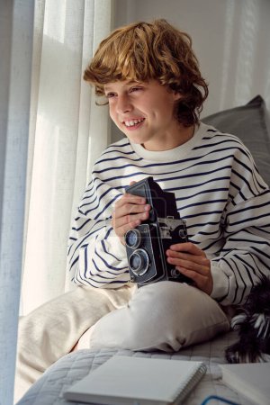 Photo for Content kid with old fashioned photo camera looking away against notebook on bed in house - Royalty Free Image