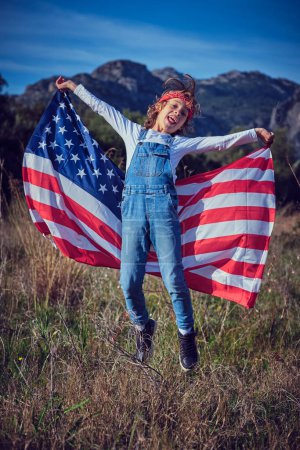 Téléchargez les photos : Corps complet de joyeux garçon préadolescent dansant et s'amusant avec le drapeau agitant des États-Unis dans prairie herbeuse - en image libre de droit
