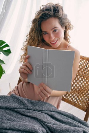 Photo for Charming young lady with long curly hair in nightie sitting on wicker chair under warm plaid near monstera plant and reading book while looking at camera - Royalty Free Image