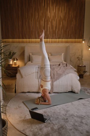 Téléchargez les photos : Jeune femme en vêtements de sport faisant du yoga dans la pose de tête soutenue dans la chambre avec ordinateur portable sur le sol - en image libre de droit