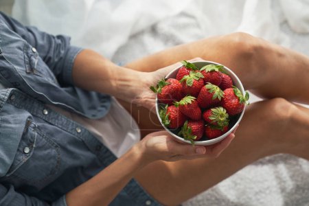 Photo for High angle of crop female holding ceramic bowl with appetizing strawberries while sitting on white fabric in light room - Royalty Free Image