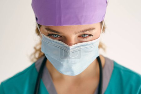 Foto de Doctora preocupada en uniforme médico con estetoscopio con máscara protectora y mirando hacia abajo sobre fondo beige - Imagen libre de derechos