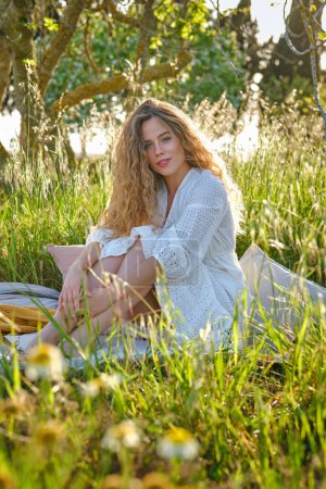 Foto de Atractiva joven hembra en elegante vestido blanco sentado en cuadros en el césped cubierto de hierba y mirando a la cámara mientras hace un picnic en el abundante jardín de verano - Imagen libre de derechos