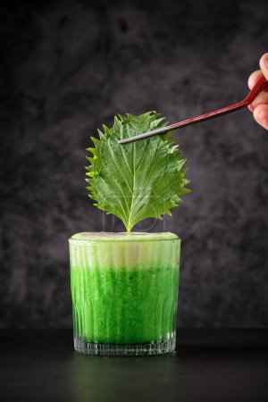 Téléchargez les photos : Barman anonyme ajoutant feuille de shiso au cocktail rafraîchissant vert avec mousse fouettée à l'aide de pinces dans le restaurant - en image libre de droit