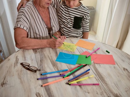 Téléchargez les photos : Recadrer anonyme grand-mère dessin sur papier coloré près du garçon tout en étant assis à la table avec tas de stylos en feutre à la maison - en image libre de droit