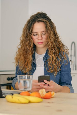 Photo for Young woman with curly hair in eyeglasses sitting at table in kitchen and browsing social media on smartphone - Royalty Free Image