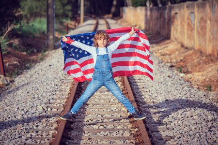 Téléchargez les photos : Corps complet de garçon préadolescent en jeans avec les mains levées et drapeau américain debout sur le chemin de fer en banlieue - en image libre de droit