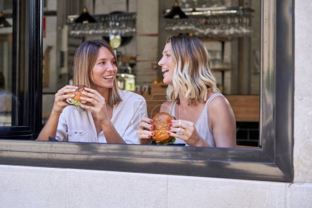 Téléchargez les photos : Délicieuses dames assises près de la fenêtre dans un café et mangeant des hamburgers savoureux tout en se regardant et en appréciant la réunion - en image libre de droit