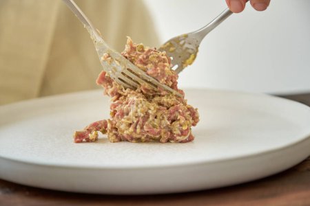 Foto de Crop cocinero sin rostro poner tartare filete en plato de servir blanco con tenedores en la mesa - Imagen libre de derechos