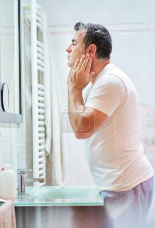 Photo for Side view of mature man wearing domestic clothes stroking face with cream during hygienic routine in bathroom while looking at himself in mirror - Royalty Free Image