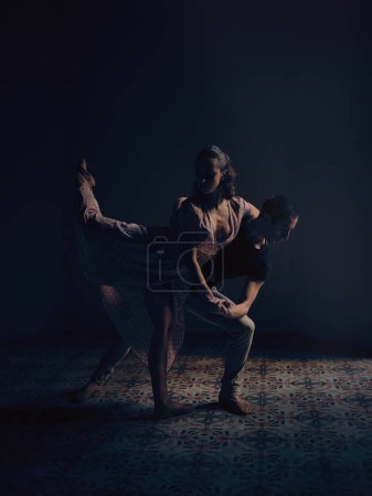 Téléchargez les photos : Vue latérale complète du corps d'une danseuse de ballet talentueuse debout avec la jambe relevée tout en s'appuyant sur l'homme dans un studio sombre - en image libre de droit
