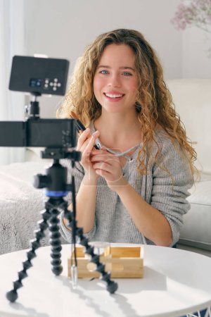 Photo for Positive young female blogger sitting at table with camera on tripod and recording video while showing makeup brushes at home - Royalty Free Image
