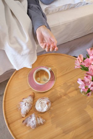 Photo for From above of anonymous woman lying in bed and reaching out round wooden table with coffee croissants and flower bouquet at bedside in morning at home - Royalty Free Image