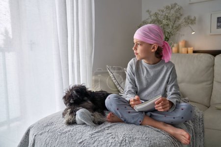 Photo for Full body of thoughtful barefoot boy in pink scarf for supporting breast cancer campaign and pajama writing wishes in notepad near Miniature Schnauzer dog lying on sofa - Royalty Free Image