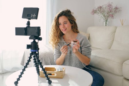 Téléchargez les photos : Une jeune blogueuse positive assise à table avec une caméra et enregistrant un tutoriel vidéo tout en montrant la palette avec des fards à paupières - en image libre de droit