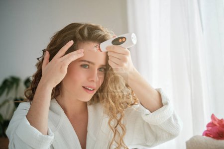 Photo for Charming female with curly hair in white bathrobe using pore remover during skincare routine while looking at reflection in mirror - Royalty Free Image