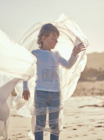 Foto de Lindo niño preadolescente de pie en la costa de arena envuelto en película de plástico transparente capturado de agua contaminada del río - Imagen libre de derechos