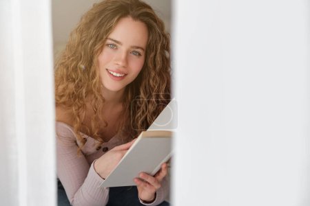 Téléchargez les photos : Charmante jeune femme aux yeux bleus lisant un roman intéressant et regardant la caméra assise près de la fenêtre avec des rideaux et se reposant à la maison - en image libre de droit