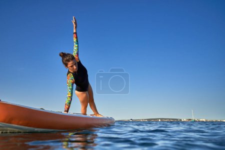 Téléchargez les photos : Corps complet de jeune athlète féminine en maillot de bain faisant Vasishthasana sur le paddleboard tout en pratiquant le yoga dans l'ondulation de la mer - en image libre de droit