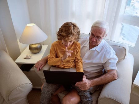 Foto de Desde arriba de hombre mayor atento con nieto navegar por Internet en netbook mientras está sentado en el sillón en la sala de estar de luz - Imagen libre de derechos